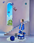 Child in a polka dot dress standing on a stool near a window with butterflies and a blue ball on the floor.