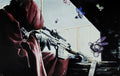 Person in a hooded cloak with a rifle, surrounded by colorful butterflies, in a dimly lit room.