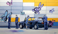Artistic depiction of a car with people and colorful butterfly-like creatures against a multicolored wall.
