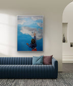 Framed artwork of a person painting a cloud on a blue sky above a modern blue sofa.