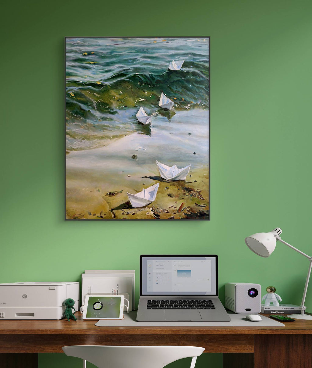 Painting of paper boats on a beach above a desk with a laptop and lamp.