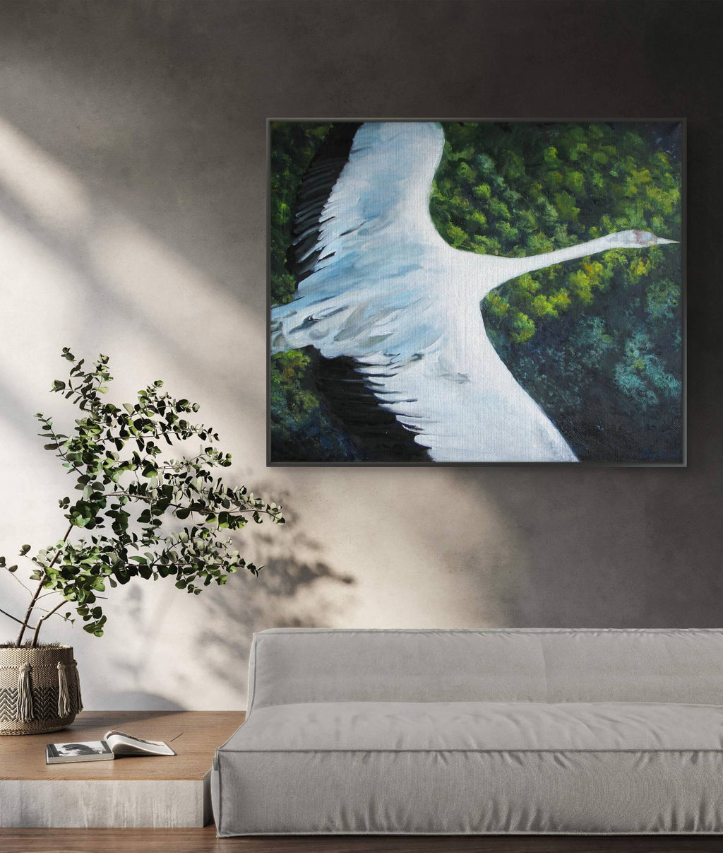 Framed painting of a swan over water with trees on a wall above a sofa and plant.