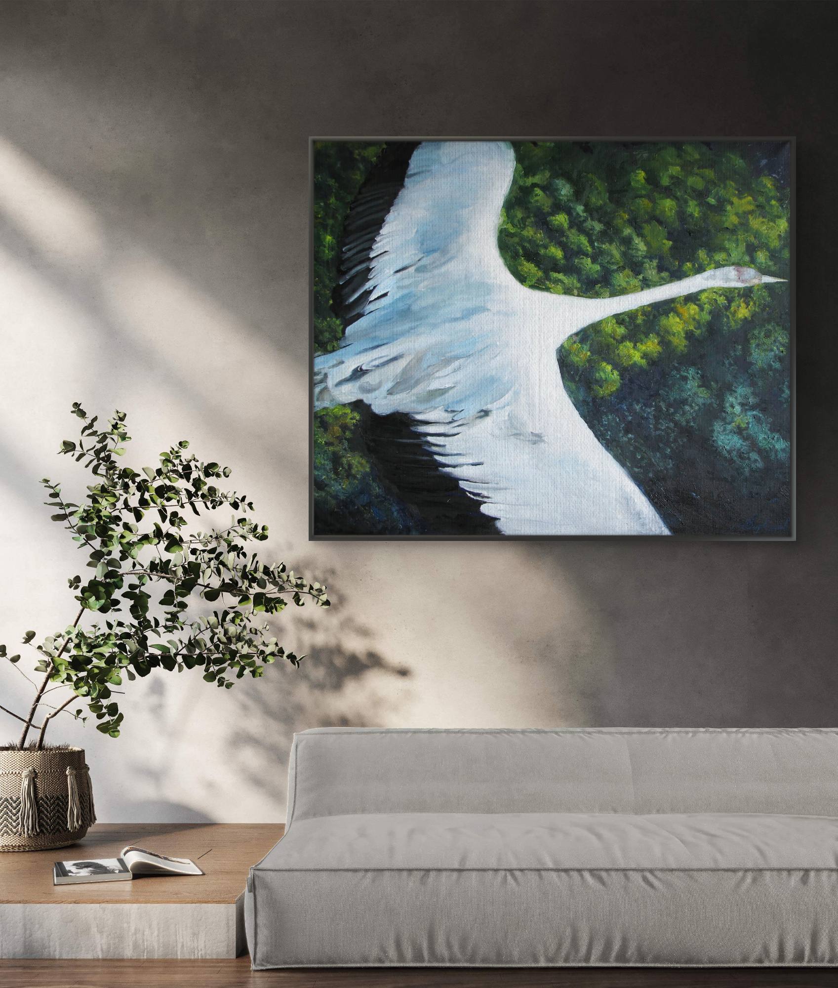 Framed painting of a swan over water with trees on a wall above a sofa and plant.