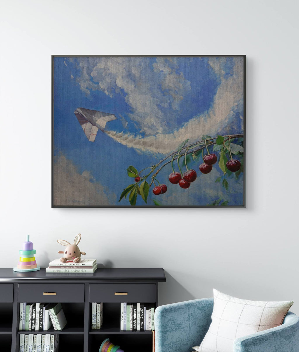 Framed artwork of a cherry branch with cherries and a plane against a blue sky with clouds, hanging on a wall above a bookshelf.
