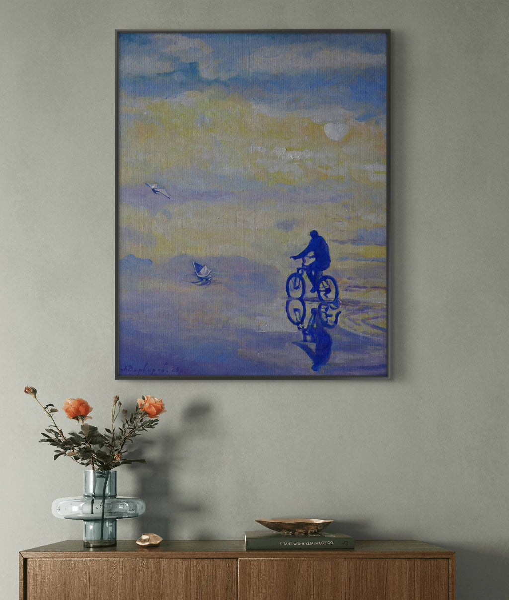 Framed artwork of a cyclist on a beach with a vase and books on a wooden surface below.