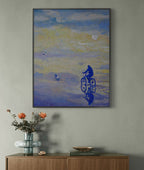 Framed artwork of a cyclist on a beach with a vase and books on a wooden surface below.